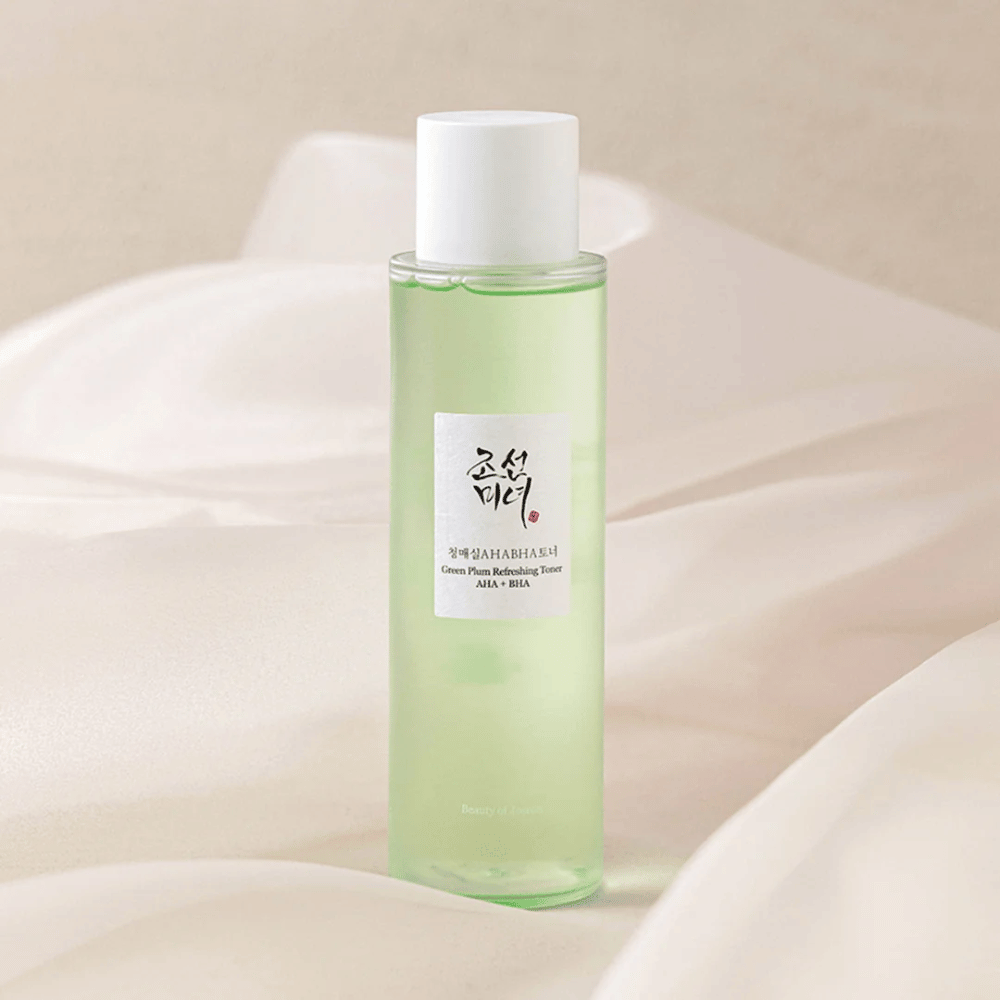 Eine Flasche mit "Green Plum Refreshing Toner" AHA + BHA steht auf einem weichen, hellen Hintergrund. Das Produkt enthält eine grüne Flüssigkeit, die Frische und Natürlichkeit widerspiegelt. Das Etikett ist weiß mit schwarzen und roten Akzenten, was einen eleganten und sauberen Look verleiht.
