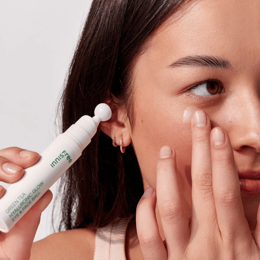Eine Person trägt Innisfree Green Tea Hyaluronic Glow Eye & Face Ball auf ihre Haut unter den Augen auf. Das Produkt hat einen weißen, runden Applikator und befindet sich in einer weißen Flasche mit grünem Text. Das Gesicht des Benutzers ist teilweise sichtbar und der Fokus liegt auf der Anwendung des Produkts.