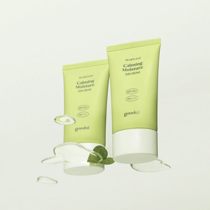 Två tuber av Goodal Heartleaf Calming Moisture Sun Cream SPF50+ PA++++ omgivna av växtblad och serumdroppar mot ljus bakgrund.