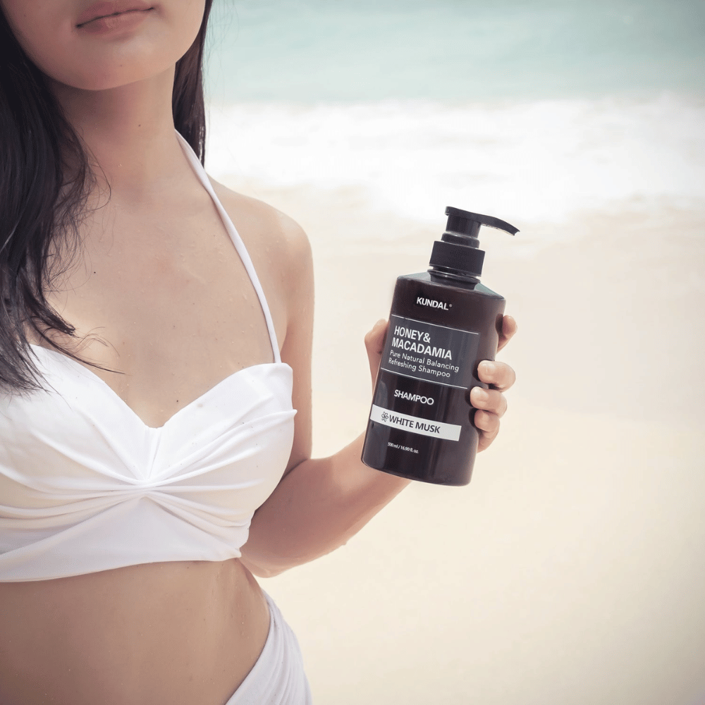 Frau im weißen Bikini hält KUNDAL Honey & Macadamia Nature Shampoo White Musk an einem Strand mit dem Meer im Hintergrund.