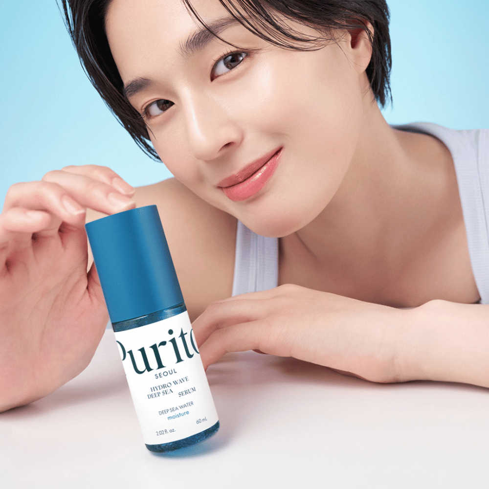 Eine Frau mit kurzem Haar hält ein blaues Serum von Purito, Hydro Wave Deep Sea Serum. Ihre Haut sieht strahlend und feucht aus, was das Produkt in den Fokus rückt. Der Hintergrund ist hellblau, was ein frisches und sauberes Gefühl vermittelt. Das Produkt ist auf Hautpflege spezialisiert.