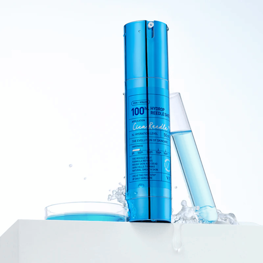 Blaue Flasche VT Cica Hyalon Hydrop Reedle Shot 100hL stehend auf weißer Fläche mit Wasserspritzern, neben einem Reagenzglas gefüllt mit blauer Flüssigkeit und einem runden Deckel mit derselben Flüssigkeit.