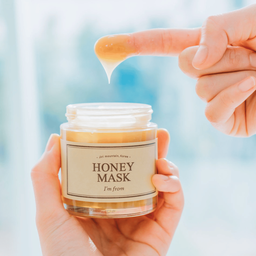 Eine Hand hält ein offenes Glas von "I'm from Honey Mask", und ein Finger wird in die goldene Maske gegen einen hellen Hintergrund getaucht.