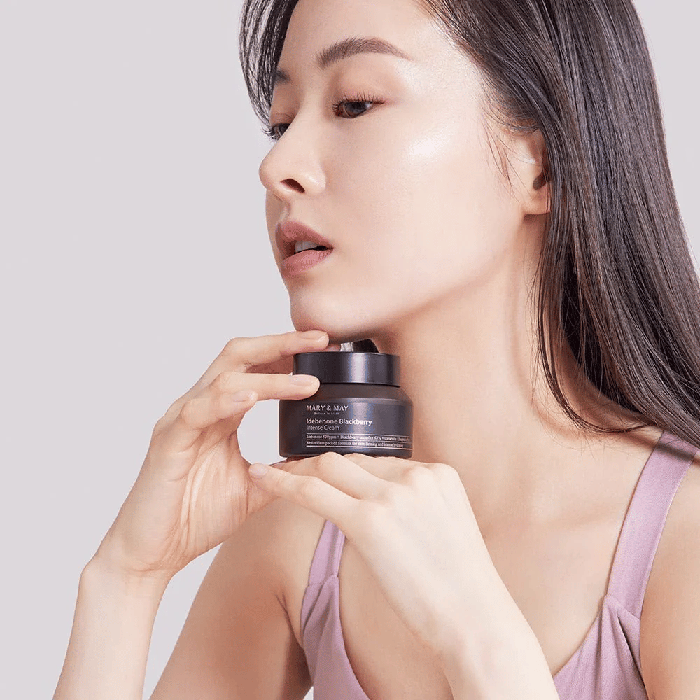 Eine Frau hält ein Glas mit MARY & MAY Idebenone Blackberry Intense Cream. Sie hat einen strahlenden Teint und lächelt leicht, gekleidet in einem ärmellosen Top. Das Produkt ist eine intensive Creme mit Idebenon und Brombeerkomplex, die für die Festigkeit und Feuchtigkeit der Haut bestimmt ist.