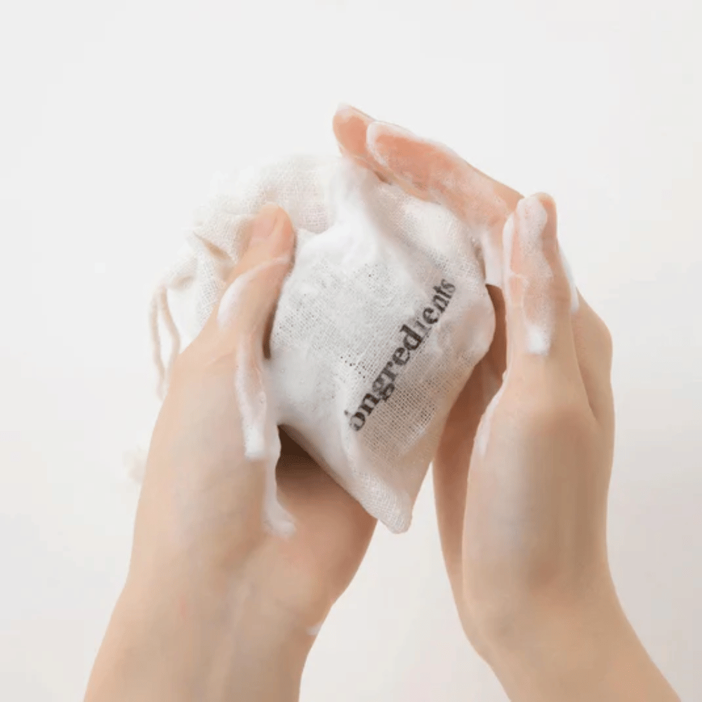 Hände, die Óngredients Jeju Green Tea Cleansing Ball in einem Stoffbeutel aufschäumen – vegane Gesichtsreinigung mit grünem Tee.