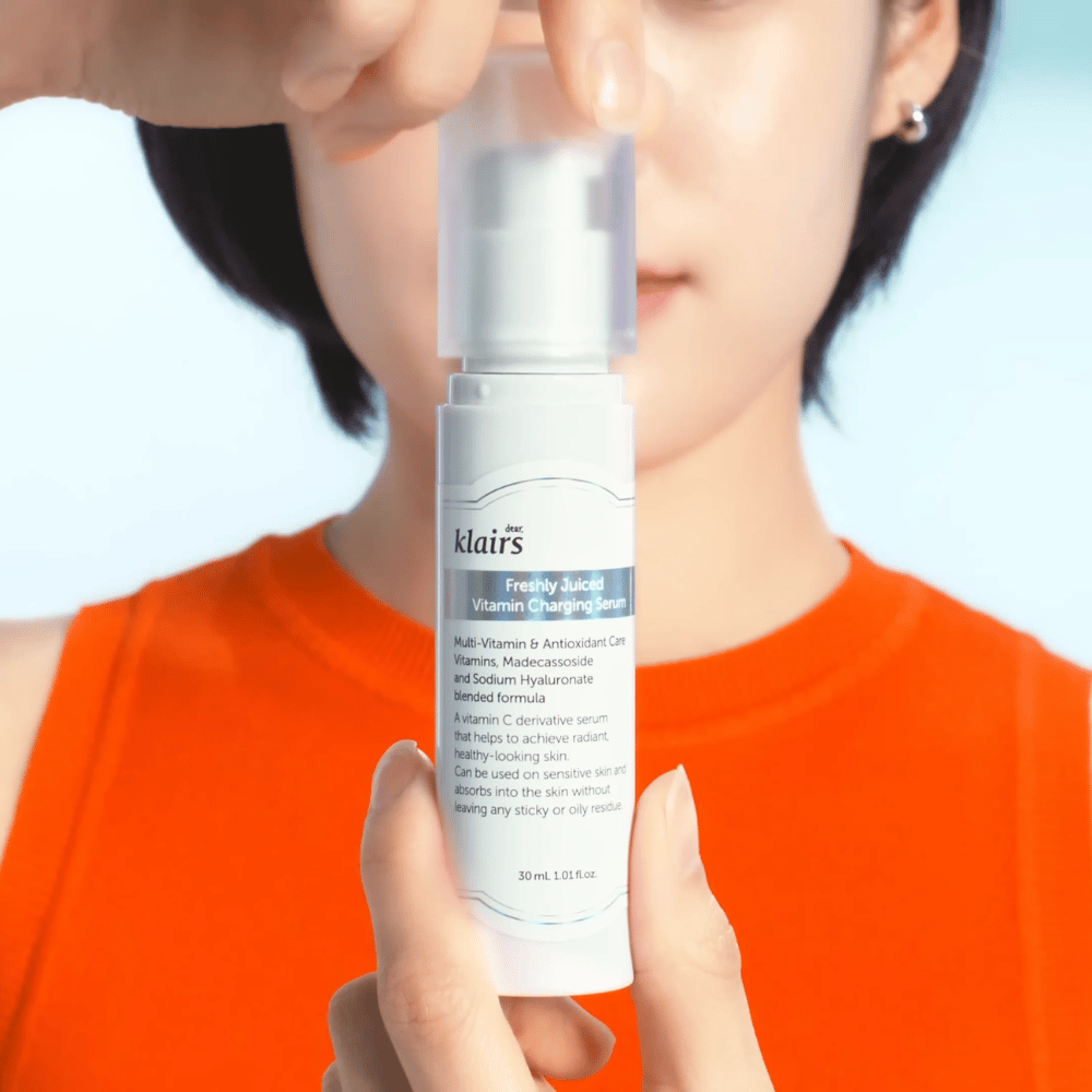 Frau im orangefarbenen Pullover hält Klairs Freshly Juiced Vitamin Charging Serum vor sich gegen einen blauen Hintergrund.