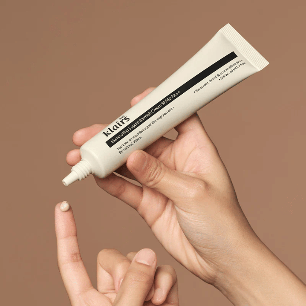 Eine Hand hält eine Tube von Klairs Illuminating Supple Blemish Cream SPF40 PA++ und drückt eine kleine Menge Creme auf einen Finger, vor einem braunen Hintergrund.