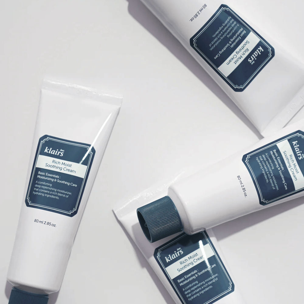 Mehrere Tuben von Klairs Rich Moist Soothing Cream strategisch auf einer hellen Fläche platziert, mit Schatten, die Tiefe erzeugen.