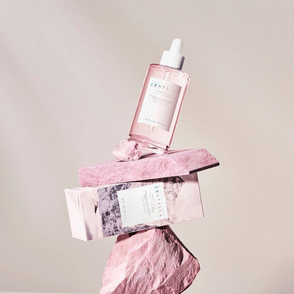 Durchsichtige Flasche mit rosa Centella-Ampulle von SKIN1004, balanciert auf einem Steinblock, mit einer Kartonverpackung darunter, vor einem neutralen Hintergrund.