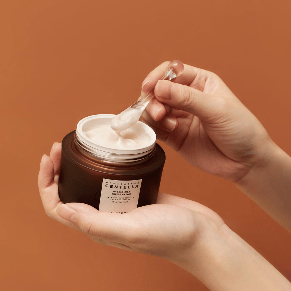 Hände, die eine offene Dose von Madagascar Centella Probio-Cica Enrich Cream und einen Spatel mit Creme vor einem einheitlich terrakottafarbenen Hintergrund halten.