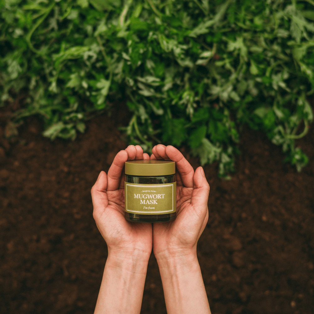 Hände, die I'm From Mugwort Mask vor einem Hintergrund aus Erde und frischen grünen Kräutern halten, was die natürlichen Inhaltsstoffe des Produkts hervorhebt.