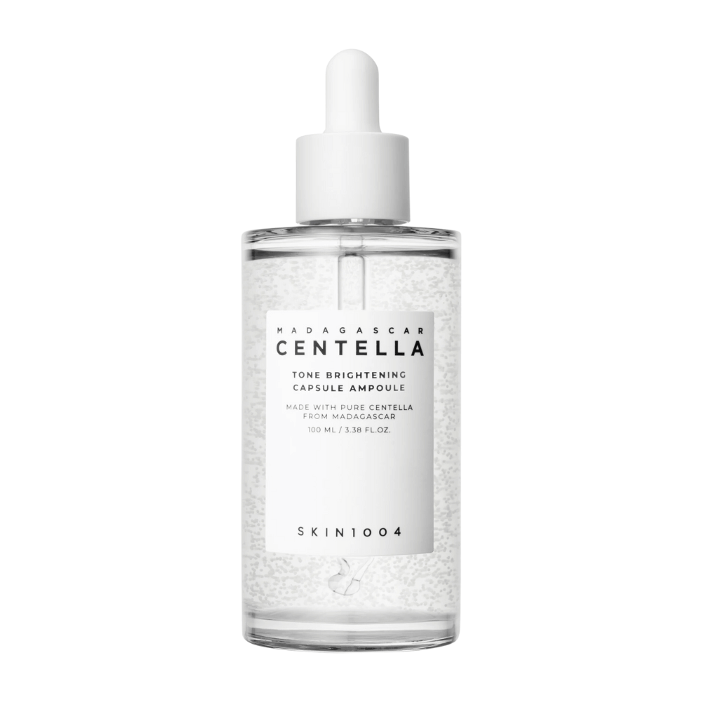 Das Bild zeigt eine Flasche von "SKIN1004 Madagascar Centella Tone Brightening Capsule Ampoule". Es ist eine durchsichtige Flasche mit einem weißen Tropfapplikator. Auf dem Etikett steht, dass das Produkt reine Centella-Inhaltsstoffe aus Madagaskar enthält. Der Inhalt der Flasche scheint kleine Kapseln oder Partikel zu enthalten, die Teil ihrer aufhellenden Hautpflegeeigenschaften sein könnten. Die Flasche enthält 100 ml / 3,38 fl.oz. des Produkts.