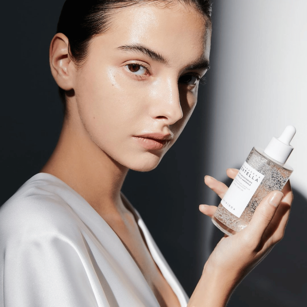 Auf dem Bild ist eine Person zu sehen, die eine Flasche mit Tropfapplikator hält. Die Flasche ist mit "SKIN1004 Madagascar Centella Tone Brightening Capsule Ampoule" gekennzeichnet. Die Person hat einen schimmernden Hautton, der das Licht reflektiert, was auf eine mögliche Wirkung des Produkts hindeutet.