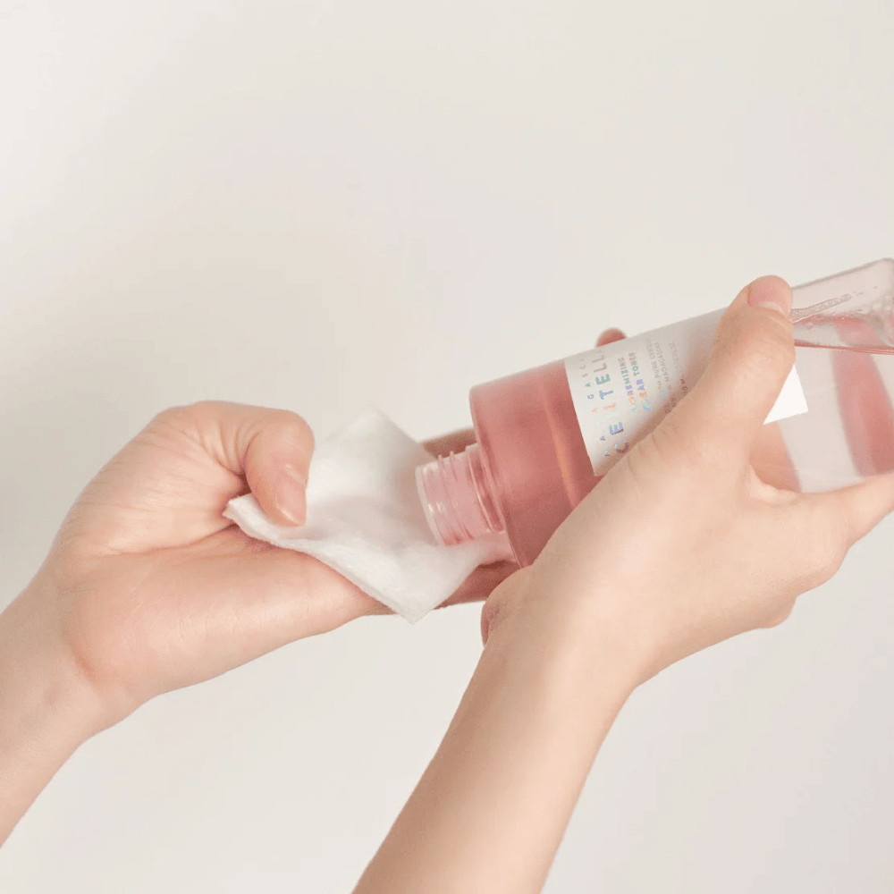 Eine Person gießt Madagascar Centella Poremizing Clear Toner aus einer transparenten Flasche auf ein Wattepad. Das Produkt, das einen rosa Farbton hat, wird verwendet, um die Haut aufzufrischen und zu reinigen. Die Hände sind sichtbar und zeigen die Anwendung des Toners.
