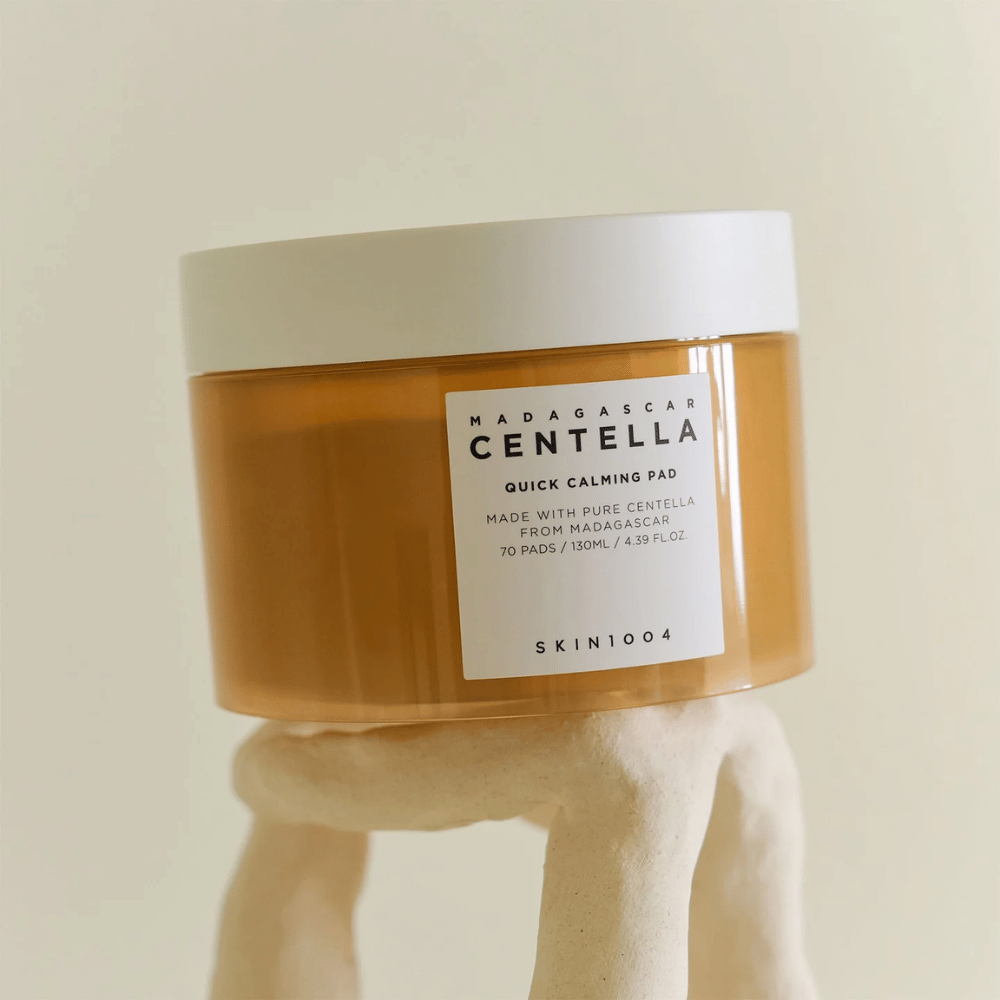 Dose mit SKIN1004 Madagascar Centella Quick Calming Pad auf einer skulpturartigen weißen Säule platziert – bernsteinfarbener Behälter mit weißem Etikett und Deckel.