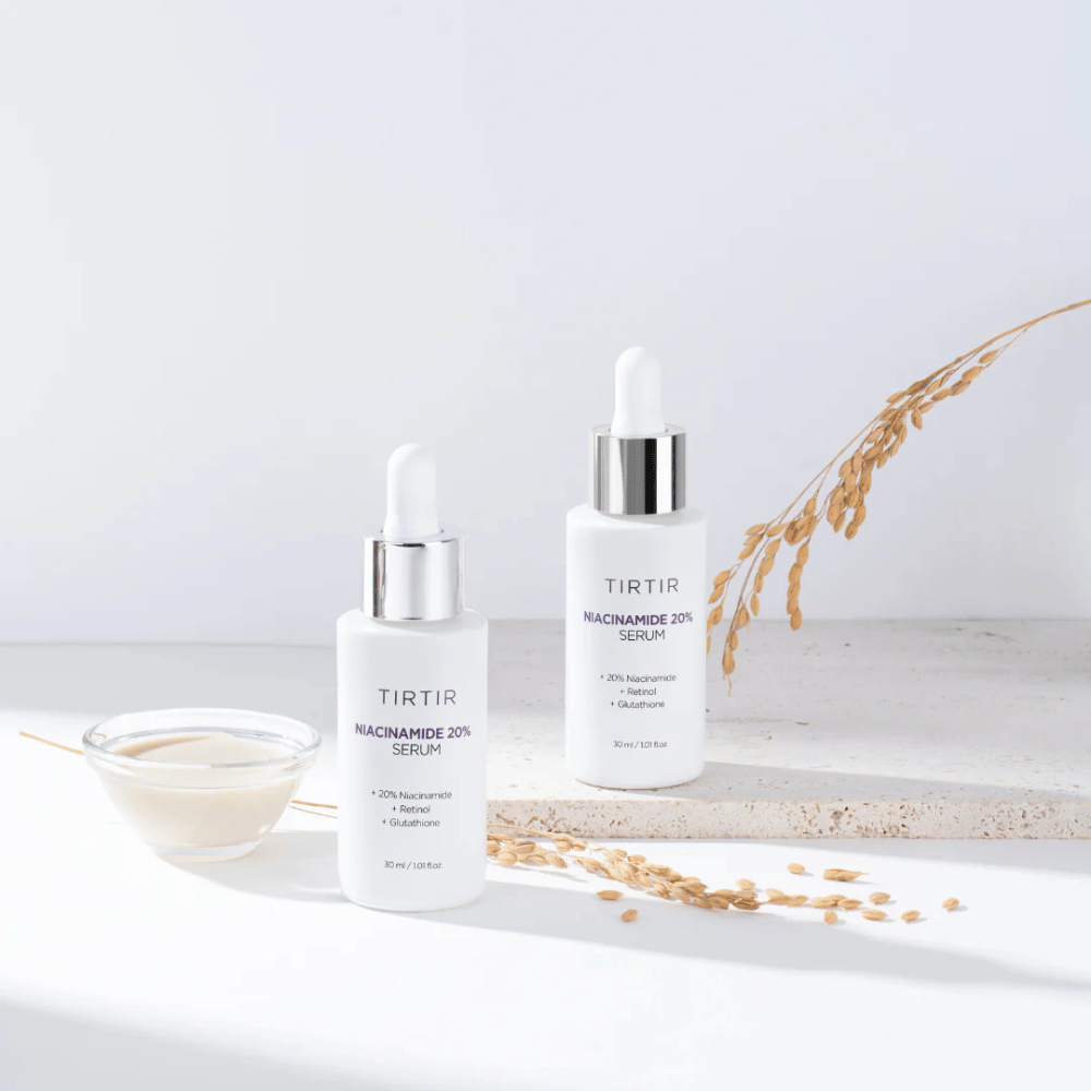 Zwei Flaschen von Tirtirs "Niacinamide 20% Serum" werden auf einer hellen Oberfläche zusammen mit einer kleinen Schale mit einer cremigen Substanz und einigen trockenen Pflanzensträngen gezeigt. Das Serum enthält 20 % Niacinamid, Retinol und Glutathion, was dazu beiträgt, das Aussehen und die Textur der Haut zu verbessern.