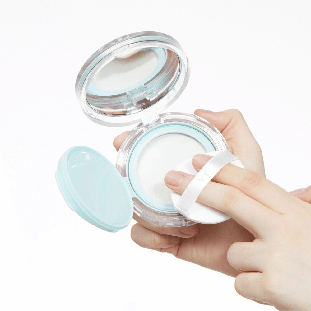 Eine Hand hält ein offenes Glas mit VT PDRN Essence Glow Sun Pact. Der Puff ist dabei, das Puder aufzutragen, das eine weiße Oberfläche und einen blauen Rand um das Glas hat.
