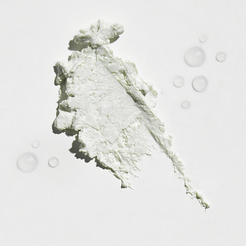 Nahaufnahme einer weißen, cremigen Substanz auf einem weißen Hintergrund, mit einigen Wassertropfen darum herum. Die Textur sieht aus wie ein Sonnenpuder oder foundation mit einem leichten Glanz.