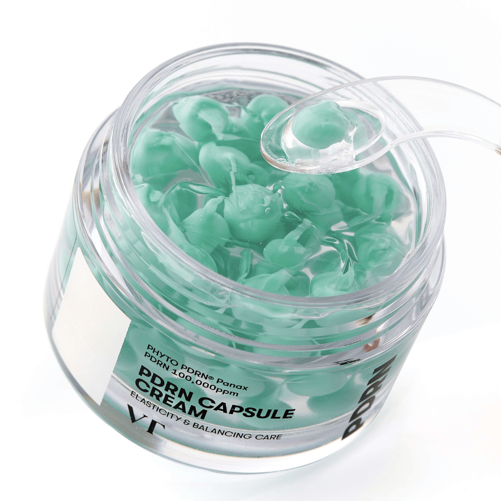 Nahaufnahme eines offenen Tiegels mit VT PDRN Kapselcreme. Der Inhalt besteht aus kleinen grünen Kapseln in einem durchsichtigen Gel, mit einem beiliegenden Spatel zur Anwendung. Der Text auf dem Tiegel lautet "Elasticity & Balancing Care".