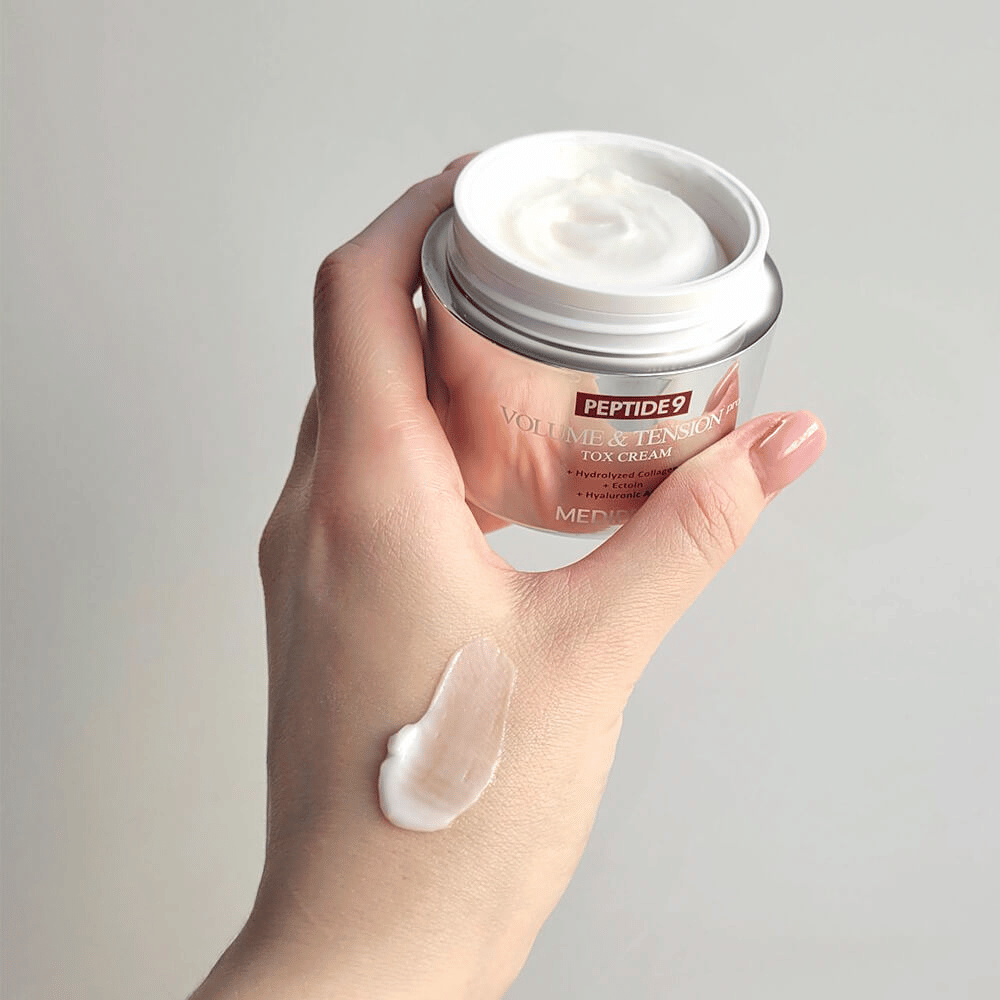 Hand håller i MEDI-PEEL Peptide 9 Volume & Tension Tox Cream med en klick kräm på handryggen. Den silkeslena formulan återfuktar och ger huden fasthet och spänst.