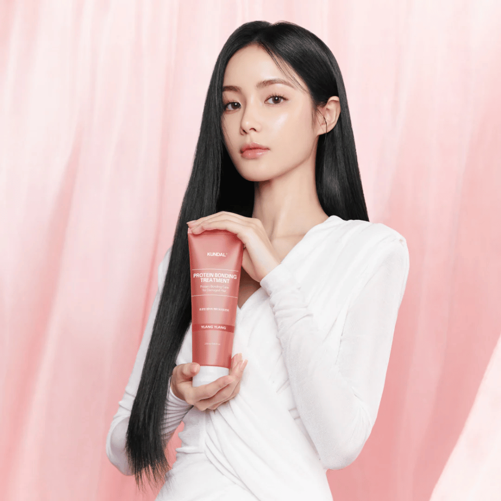 Frau mit langem, glänzendem Haar hält KUNDAL Protein Bonding Treatment Violet Muguet vor rosa Hintergrund.