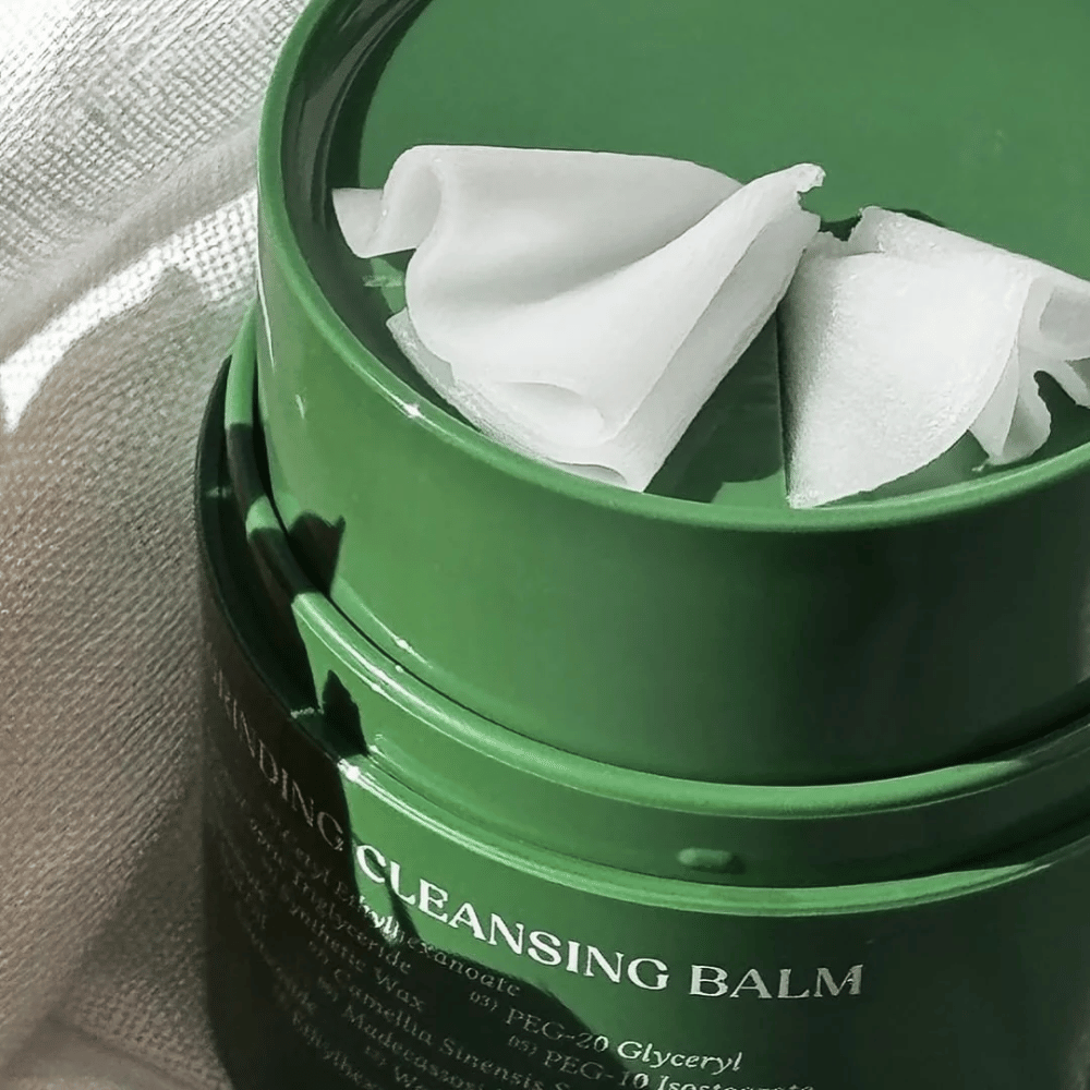 Das Bild zeigt eine grüne Dose mit "Cleansing Balm", die geöffnet ist, um den mitgelieferten Spatel und den weichen Inhalt zu enthüllen. Die cremige Konsistenz reflektiert das Licht, was ein Gefühl von Luxus und Effizienz bei der Reinigung vermittelt.