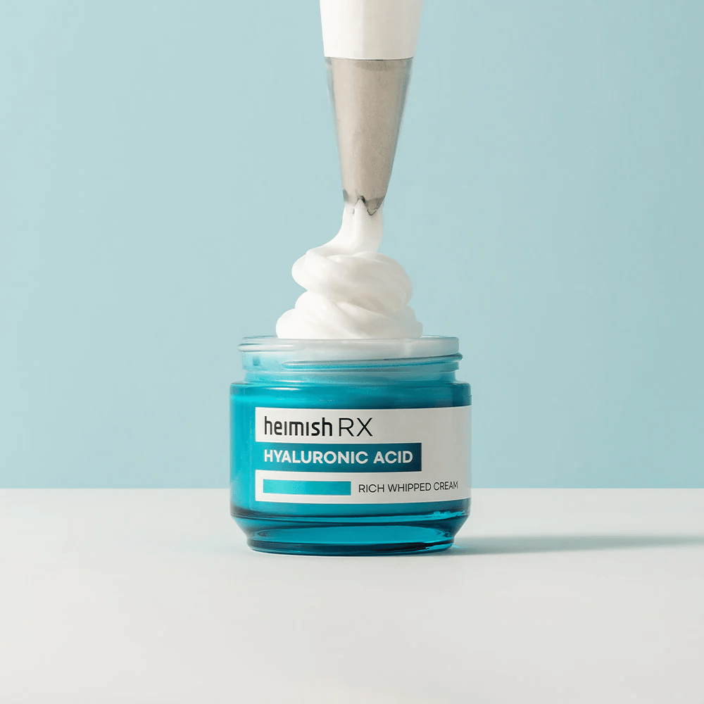 Heimish RX Hyaluronic Acid Rich Whipped Cream in einem blauen Glasgefäß, wo ein Spritzbeutel die fluffige Creme aufträgt. Eine feuchtigkeitsspendende Gesichtscreme mit Hyaluronsäure für intensive Feuchtigkeit und eine weiche, pralle Haut.
