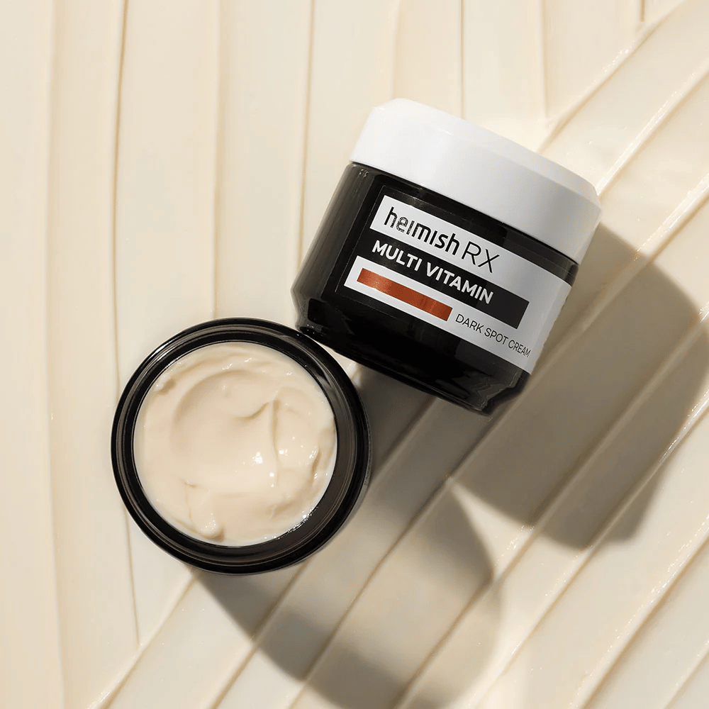 heimish RX Multi Vitamin Dark Spot Cream – eine nährstoffreiche Gesichtscreme zur Reduzierung dunkler Flecken und zur Verbesserung des Hauttons. Die cremige Textur der Creme wird in einem offenen Tiegel mit eleganter Verpackung gezeigt.