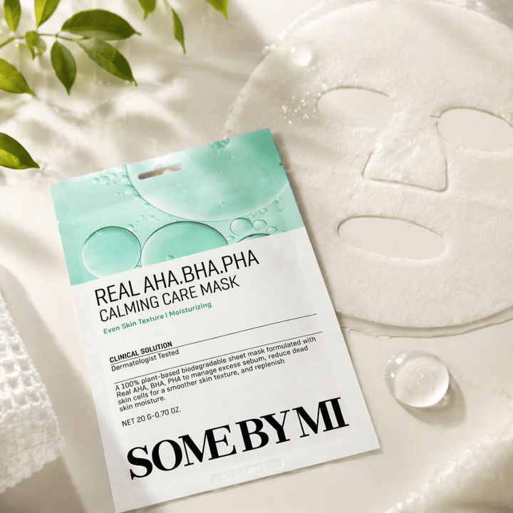 SOME BY MI Real AHA.BHA.PHA Calming Care Mask wird zusammen mit der entfalteten Tuchmaske gezeigt. Um das Produkt herum sind Feuchtigkeitstropfen und grüne Blätter zu sehen, die das Gefühl von Feuchtigkeitsversorgung, beruhigender Pflege und sanftem Peeling verstärken. Das Bild hebt den Fokus der Maske auf ein ebenmäßigeres Hautbild und eine verbesserte Feuchtigkeitsbalance hervor.