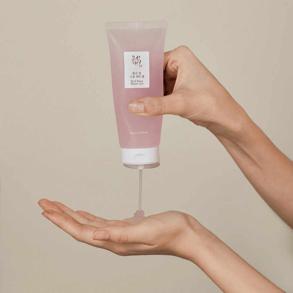 Das Bild zeigt eine Person, die eine Tube von "Red Bean Water Gel" von Beauty of Joseon hält und ein durchsichtiges Gel in ihre Hand drückt. Das Produkt scheint für die Hautpflege bestimmt zu sein und hat eine attraktive, sanft rosa Verpackung.