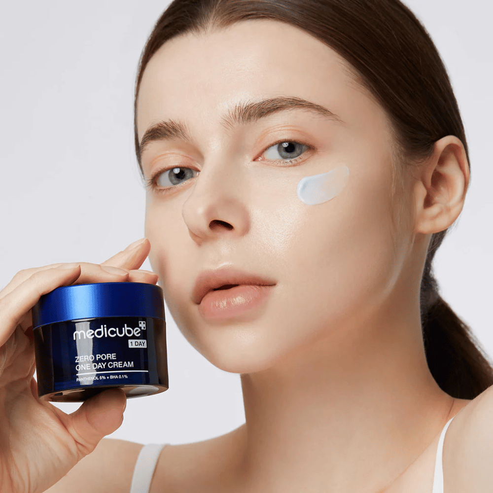 Frau mit einem natürlichen Look hält Medicube Zero Pore One Day Cream. Eine kleine Menge Creme ist auf ihrer Wange aufgetragen. Das Produkt ist darauf ausgelegt, die Hautstruktur zu verbessern und die Porengröße zu minimieren, mit einer leichten und feuchtigkeitsspendenden Formel.