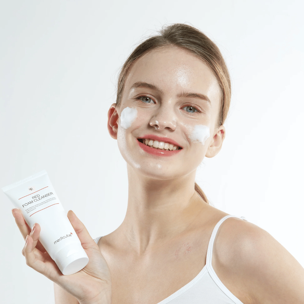 Junge Frau mit Reinigungsschaum im Gesicht hält eine Tube von Medicube Red Foam Cleanser hoch und lächelt in die Kamera. Sie trägt ein weißes Trägershirt und der Hintergrund ist hell und schlicht.