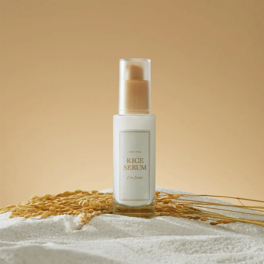 Flasche mit "I'm from" Rice Serum, umgeben von Reiskörnern und Reisähren vor einem warmen Hintergrund.