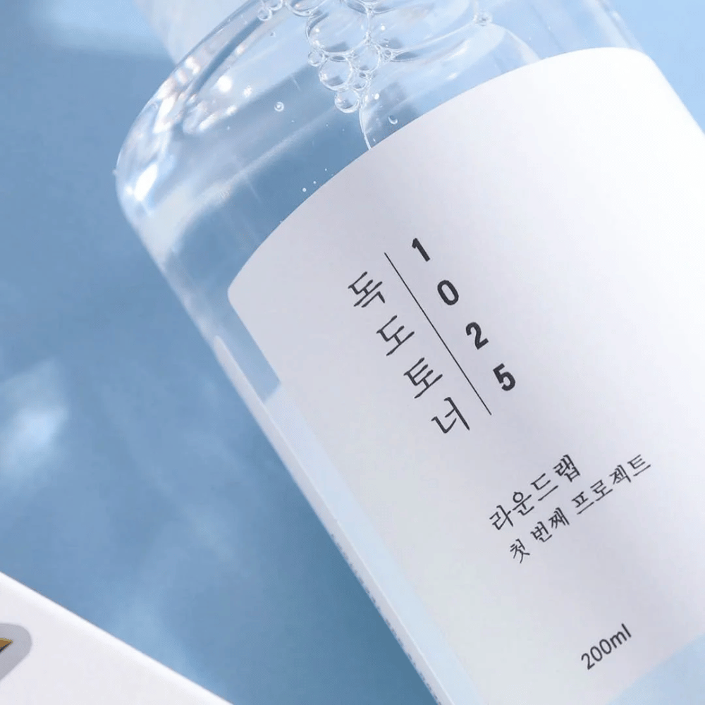 Nahaufnahme einer durchsichtigen Flasche Round Lab 1025 Dokdo Toner, gefüllt mit klarer Flüssigkeit, vor einem blauen Hintergrund.