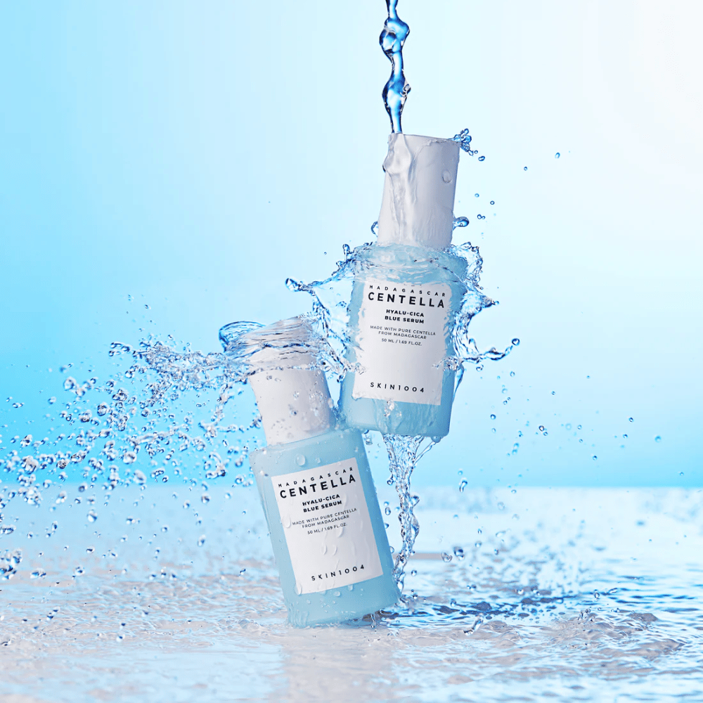 Zwei Flaschen von SKIN1004 Madagascar Centella Hyalu-Cica Blue Serum werden von einem Wasserstrahl vor einem hellblauen Hintergrund getroffen.