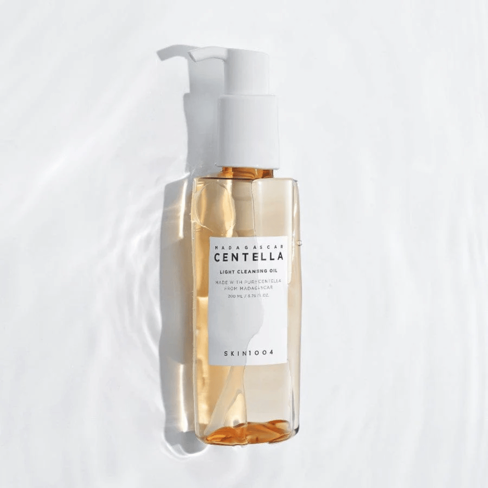 SKIN1004 Madagascar Centella Light Cleansing Oil auf weißem Hintergrund mit weichen Wasserreflexen, die die transparente Flasche und den goldenen Inhalt hervorheben.
