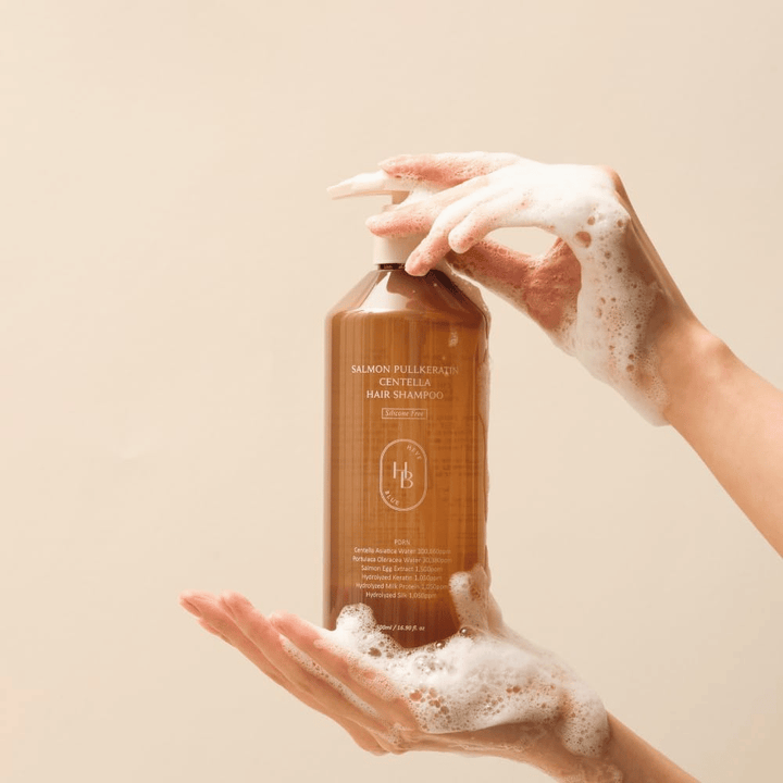 Heveblue Salmon Pullkeratin Centella Hair Shampoo in brauner Flasche, umgeben von Schaum. Das Produkt wird zwischen zwei Händen gehalten und hebt ein reinigendes und pflegendes Haarpflegeerlebnis mit reichlich Schaum hervor.