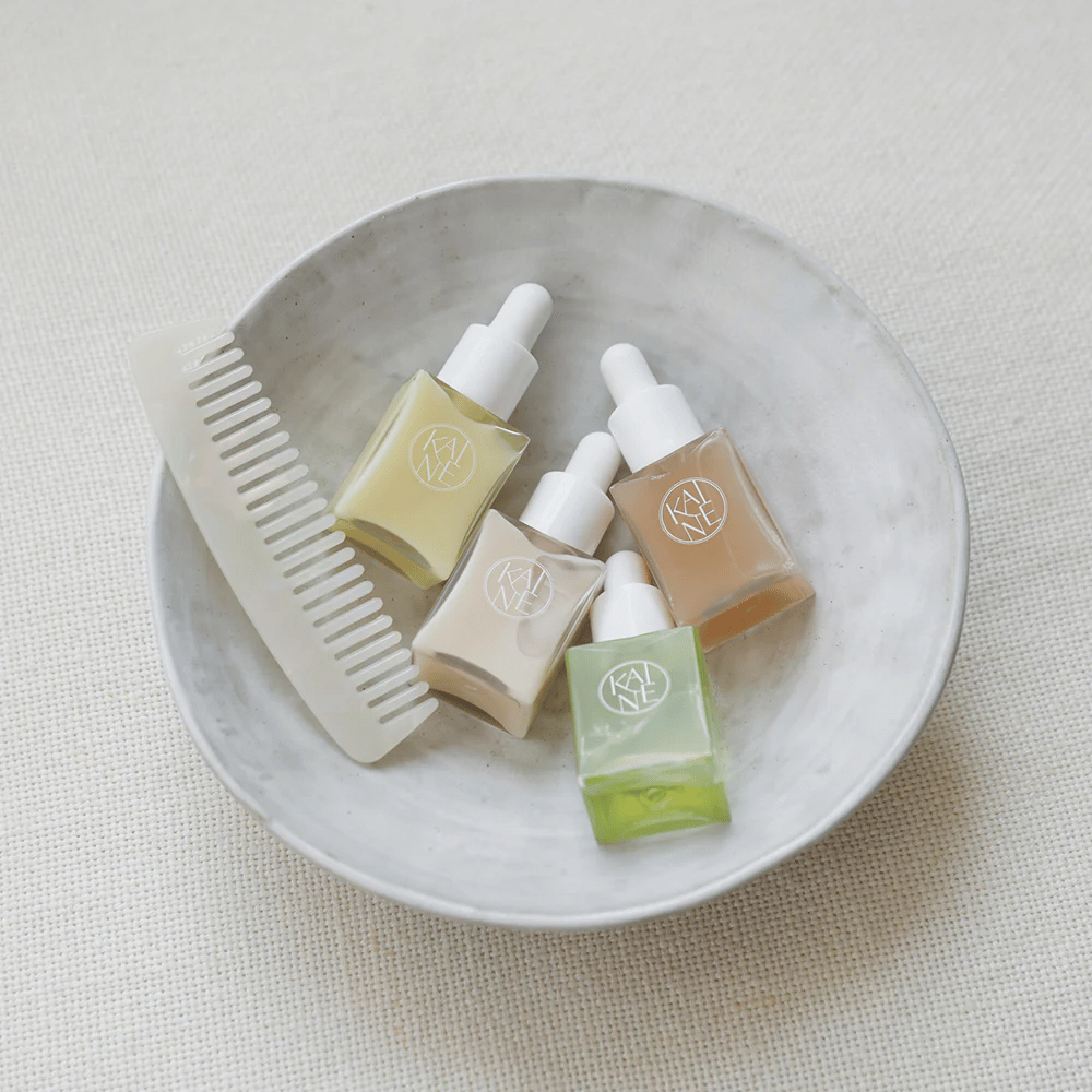 Vier kleine Serumflaschen von KAINÉ Serum Discovery Kit liegen in einer weißen Schale neben einem Kamm, mit einem natürlichen und ruhigen Hintergrund in Beigetönen.