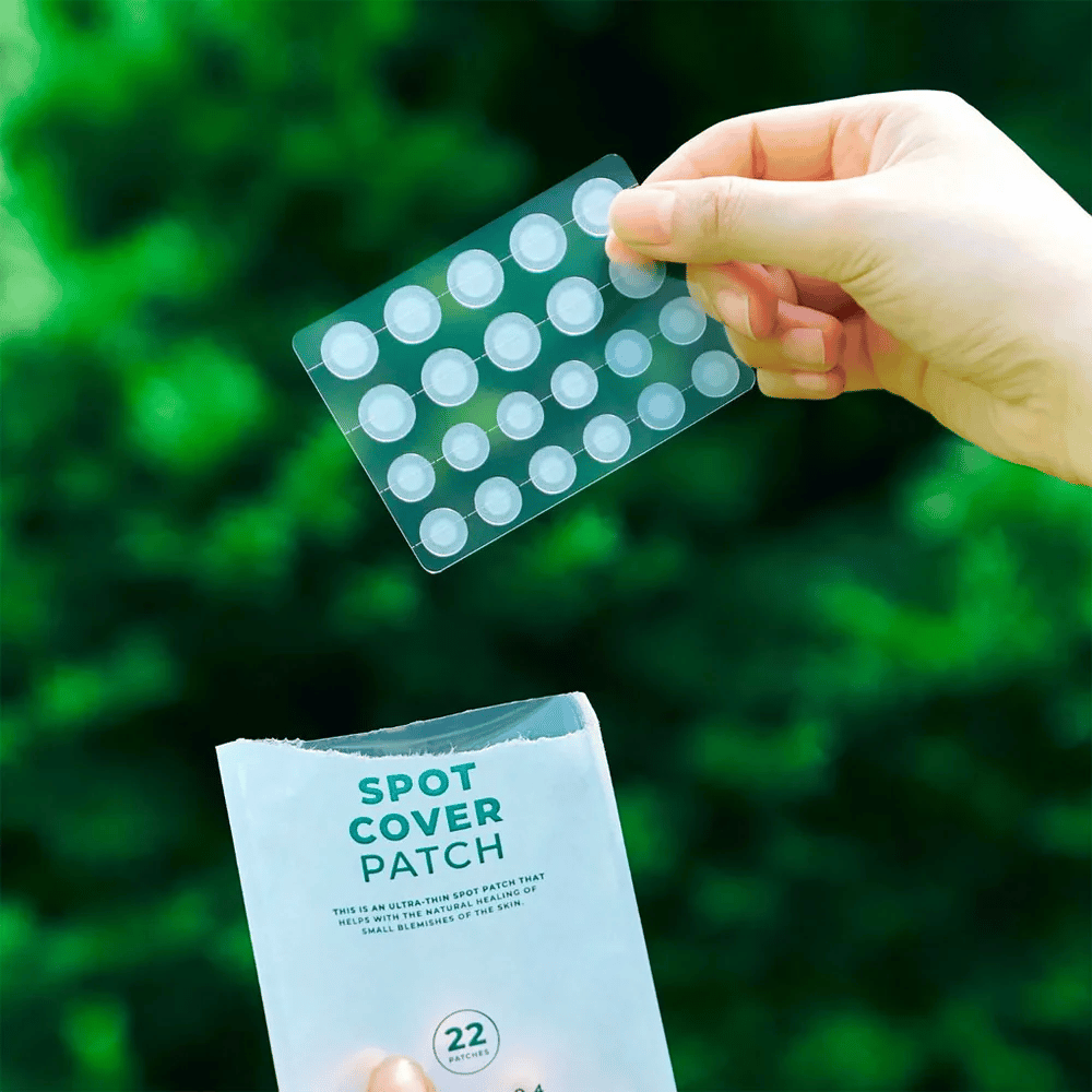 Eine Hand hält ein Blatt mit Spot Cover Patches, die transparent und abgerundet sind. Im Hintergrund ist eine verschwommene grüne Umgebung zu sehen. Die Verpackung zeigt einen Teil der Verpackung mit dem Text "SPOT COVER PATCH" und Informationen darüber, dass das Produkt 22 Pflaster enthält, die helfen, kleine Hautbereiche zu heilen.