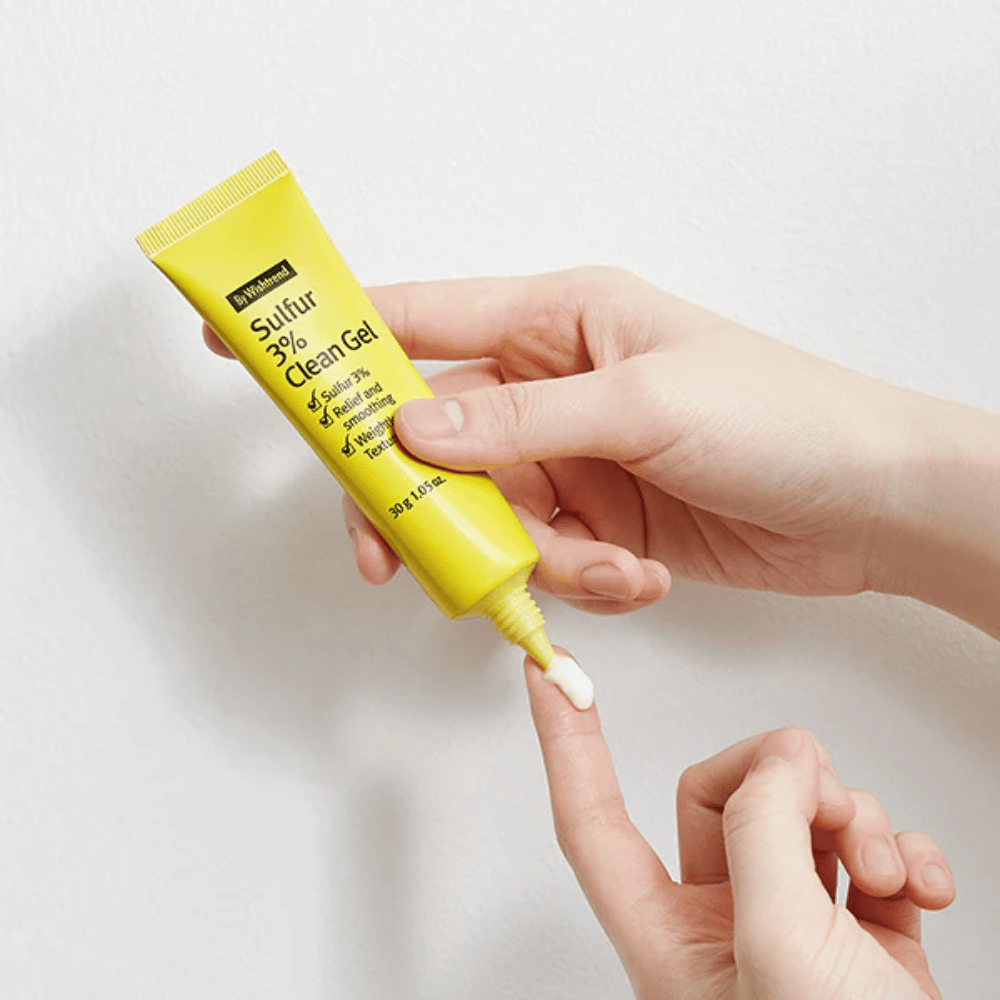 Nahaufnahme von Händen, die By Wishtrend Sulfur 3% Clean Gel auftragen, eine gelbe Tube mit weißem Gel, vor einem weißen Hintergrund.