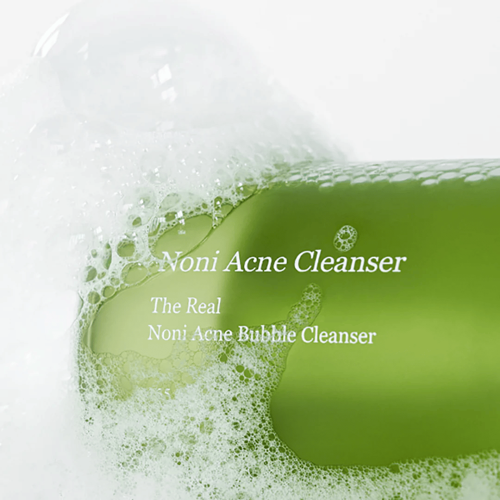 Die Flasche mit The Real Noni Acne Bubble Cleanser steht im Mittelpunkt und ist von reichlich Schaum umgeben. Die frische grüne Farbe signalisiert natürliche Inhaltsstoffe, perfekt zum Reinigen und zur Bekämpfung von Akne. Ein ideales Produkt für eine reine und klare Haut.