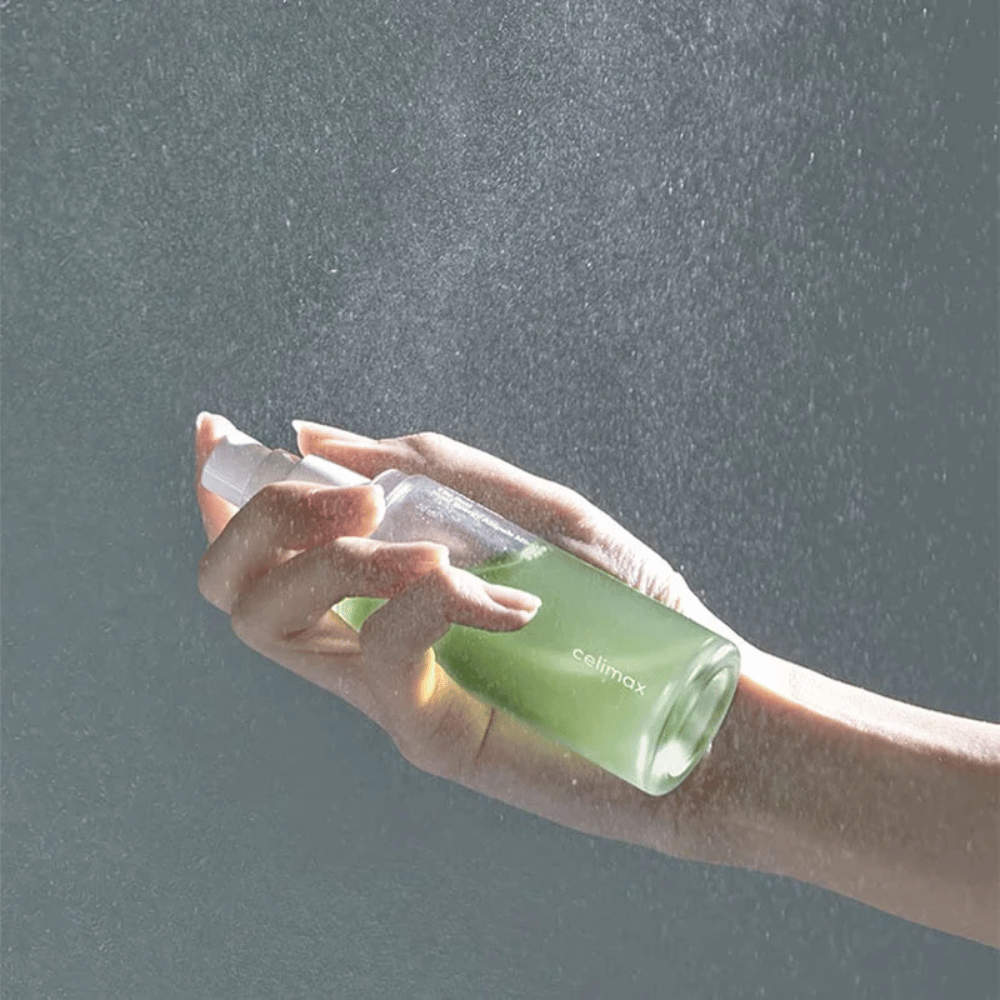 Eine Hand hält Celimax Noni Mist Sprühflasche und trägt das Produkt in einem feinen, gleichmäßigen Sprühstoß auf die Haut auf. Die Flasche ist grün und das Produkt wird als "The Real Noni Energy Ampule Mist" vermarktet, um der Haut Energie und Feuchtigkeit zu spenden.