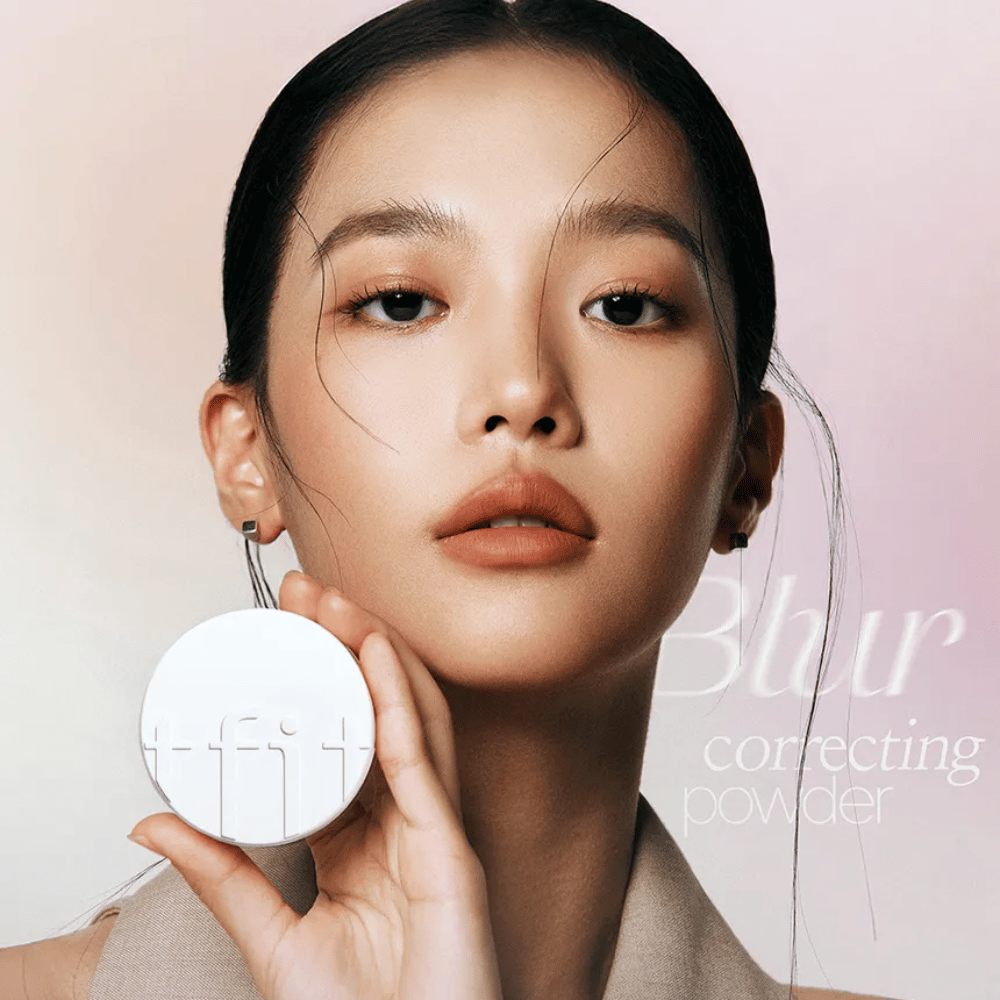 Kvinna med felfri makeup håller upp en rund vit dosa med TFIT-logga, med texten "Blur Correcting Powder" i bakgrunden.