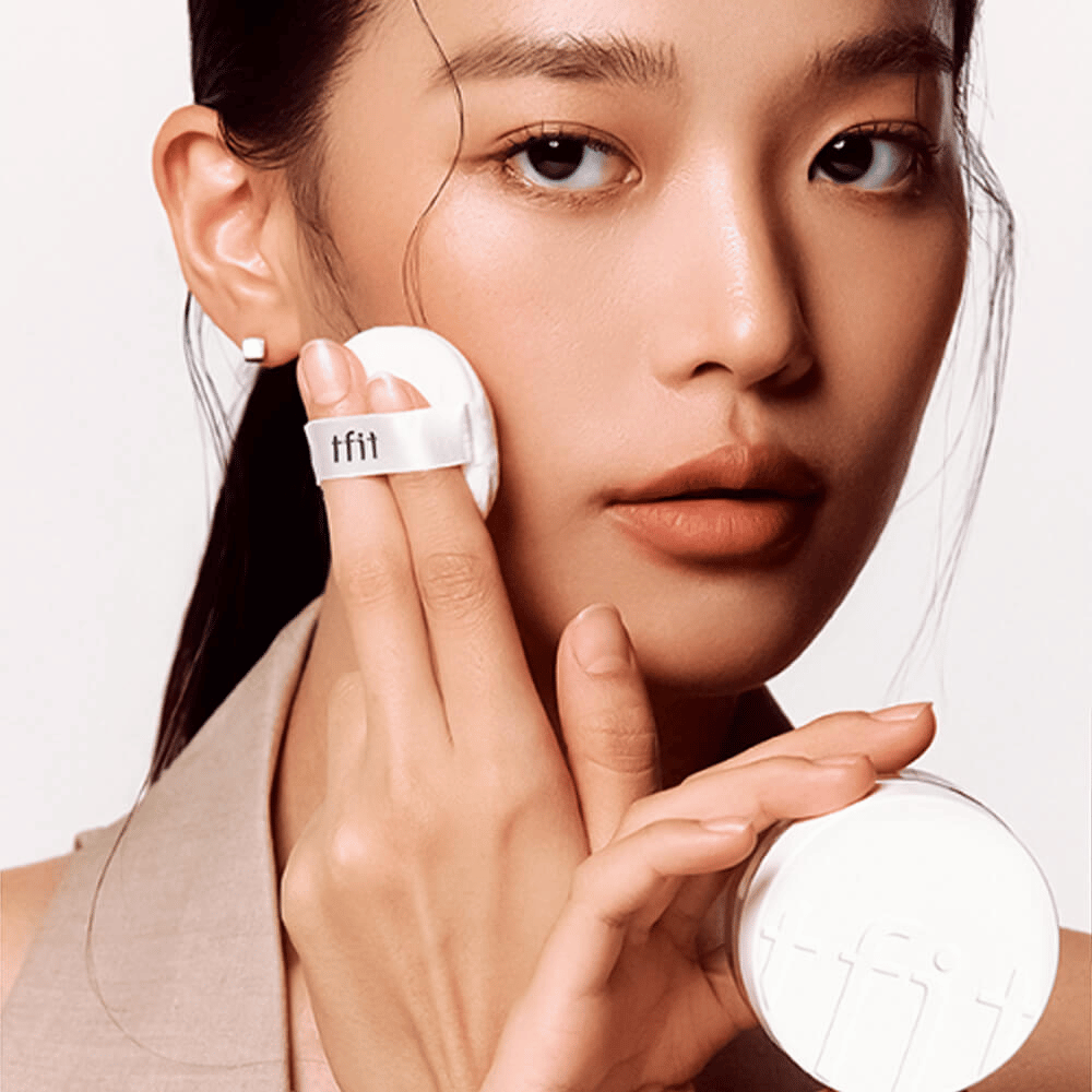 Kvinna applicerar TFIT Translucent Set Finishing Powder #02 Skin Beige med en puff, håller i en vit puderburk med TFIT-logga.