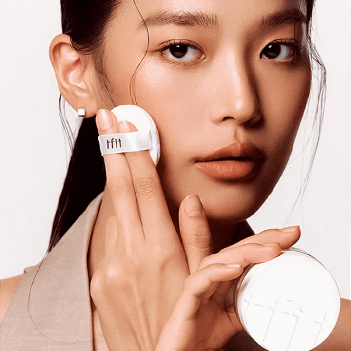 Kvinna applicerar TFIT Translucent Set Finishing Powder #02 Skin Beige med en puff, håller i en vit puderburk med TFIT-logga.