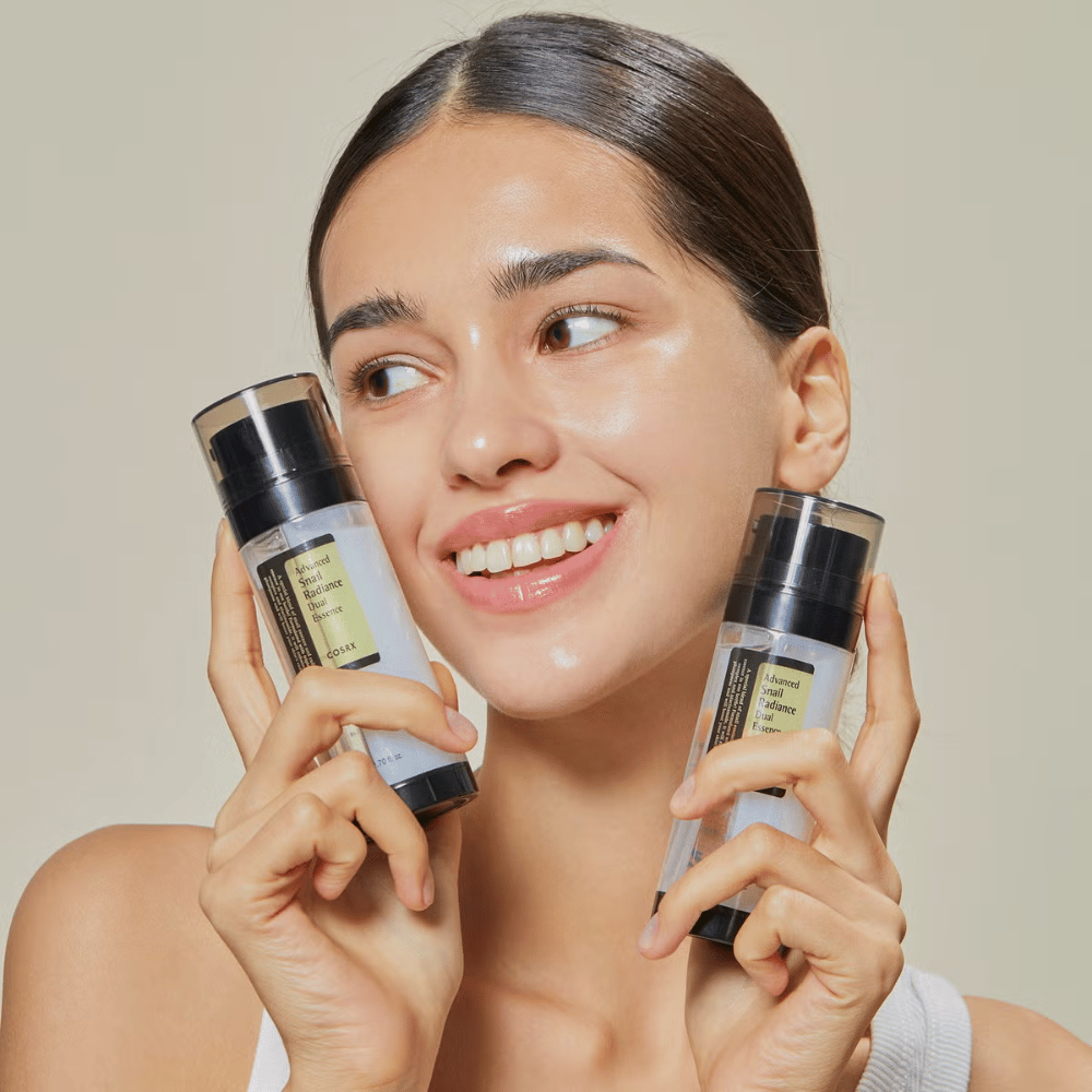 Lächelnde Frau mit glänzender Haut hält zwei Flaschen COSRX Advanced Snail Radiance Dual Essence 80 ml nahe am Gesicht vor einem hellen Hintergrund.