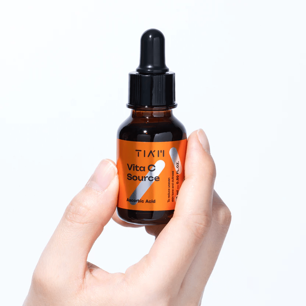 Hand hält TIA'M Vita C Source, ein Vitamin-C-Serum in einer braunen Glasflasche mit Pipette. Das Produkt ist darauf ausgelegt, den Hautton auszugleichen und Glanz zu verleihen.