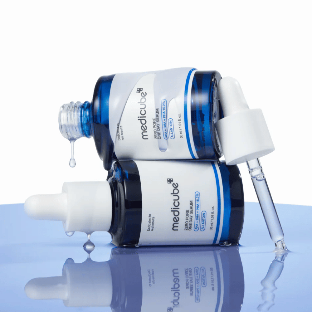 Zwei blaue Flaschen von Medicube Zero Pore One Day Serum, die gegeneinander liegen, mit Pipetten und Serumtropfen auf einer reflektierenden blauen Fläche.