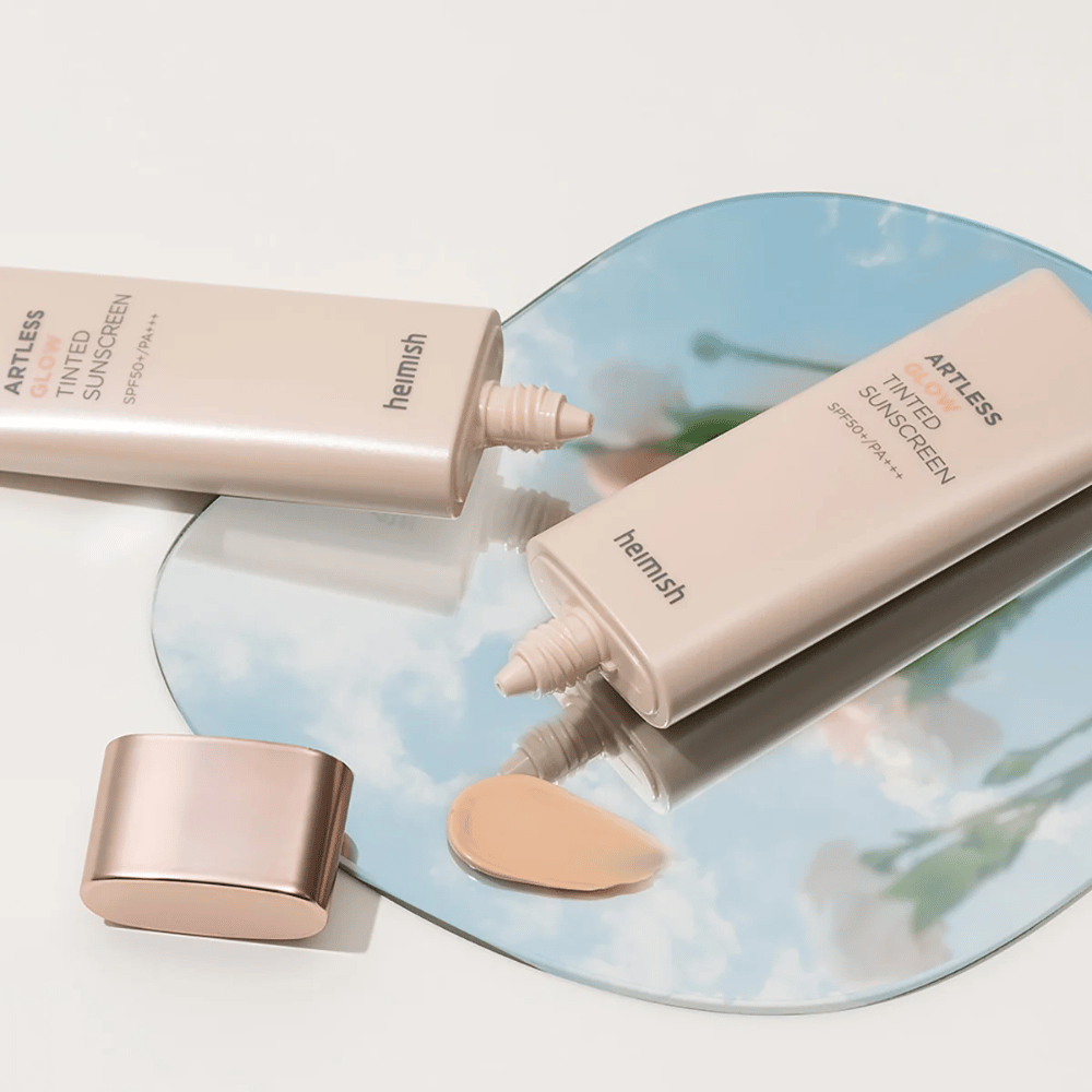 Eine Flasche heimish Artless Glow Tinted Sunscreen SPF50+ PA+++ 40ml und eine Flasche concealer auf einem Spiegel, der verschiedene Hauttöne zeigt.