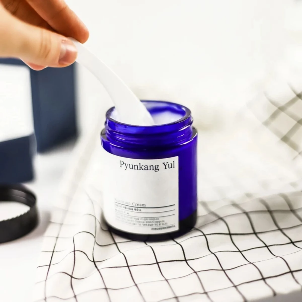 Eine Person benutzt eine Dose Pyunkang Yul Nutrition Cream 100ml auf einem Tisch.